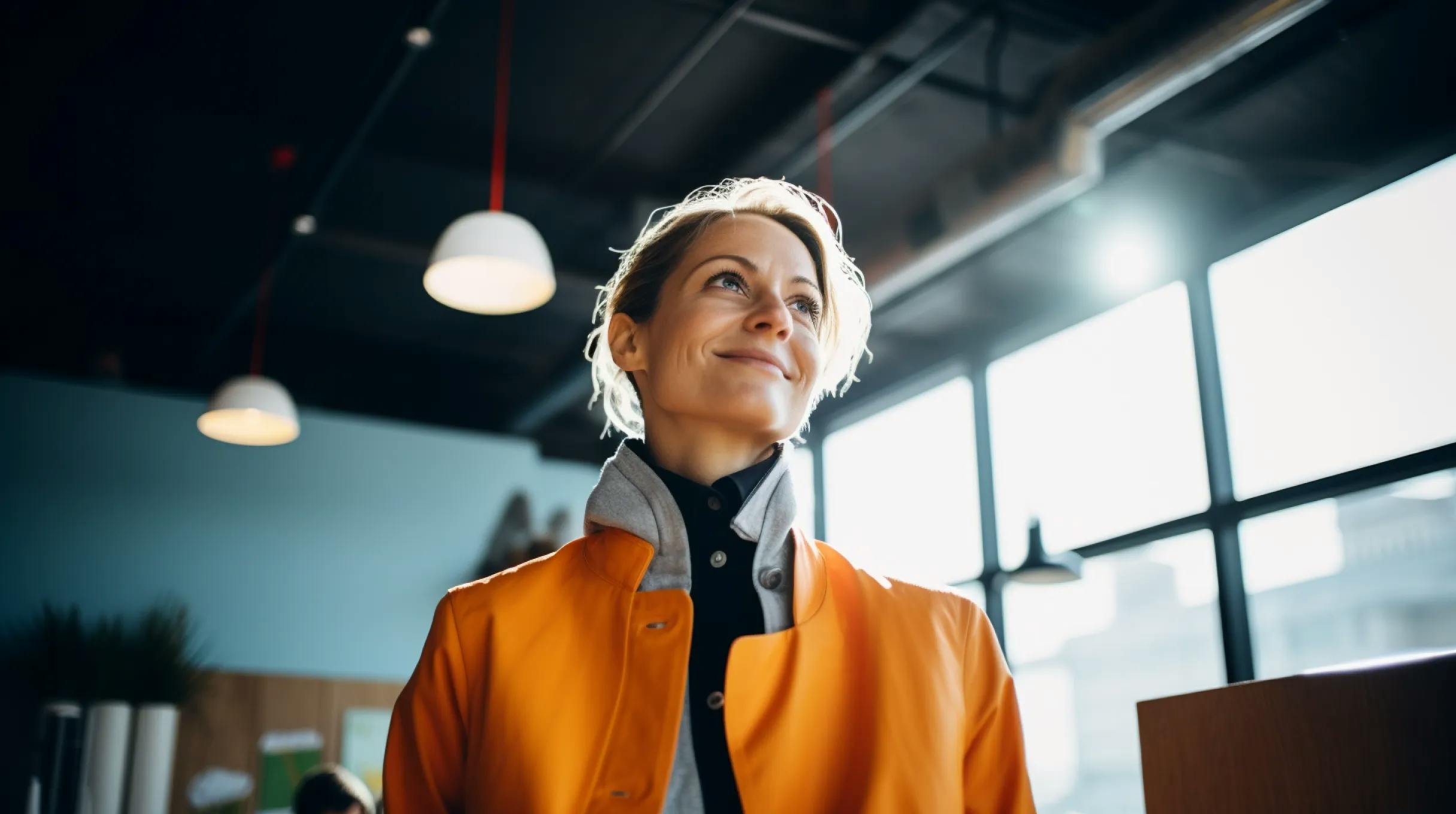  A woman in an orange jacket standing confidently in a well-lit office space.